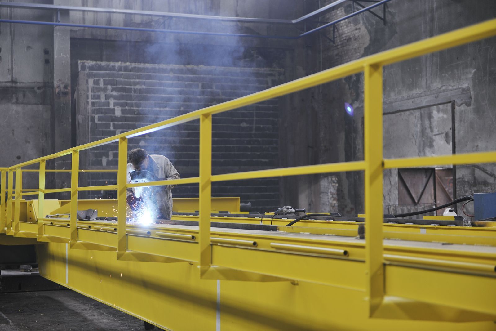 A person wearing protective gear welds on a bright yellow industrial bridge structure inside a workshop.