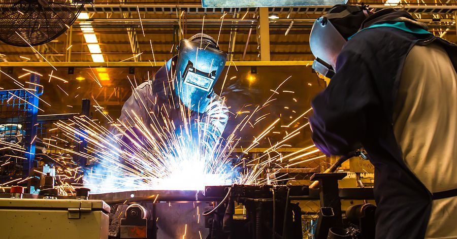 Two workers wearing protective welding helmets and gear operate in an industrial facility amid bright sparks.