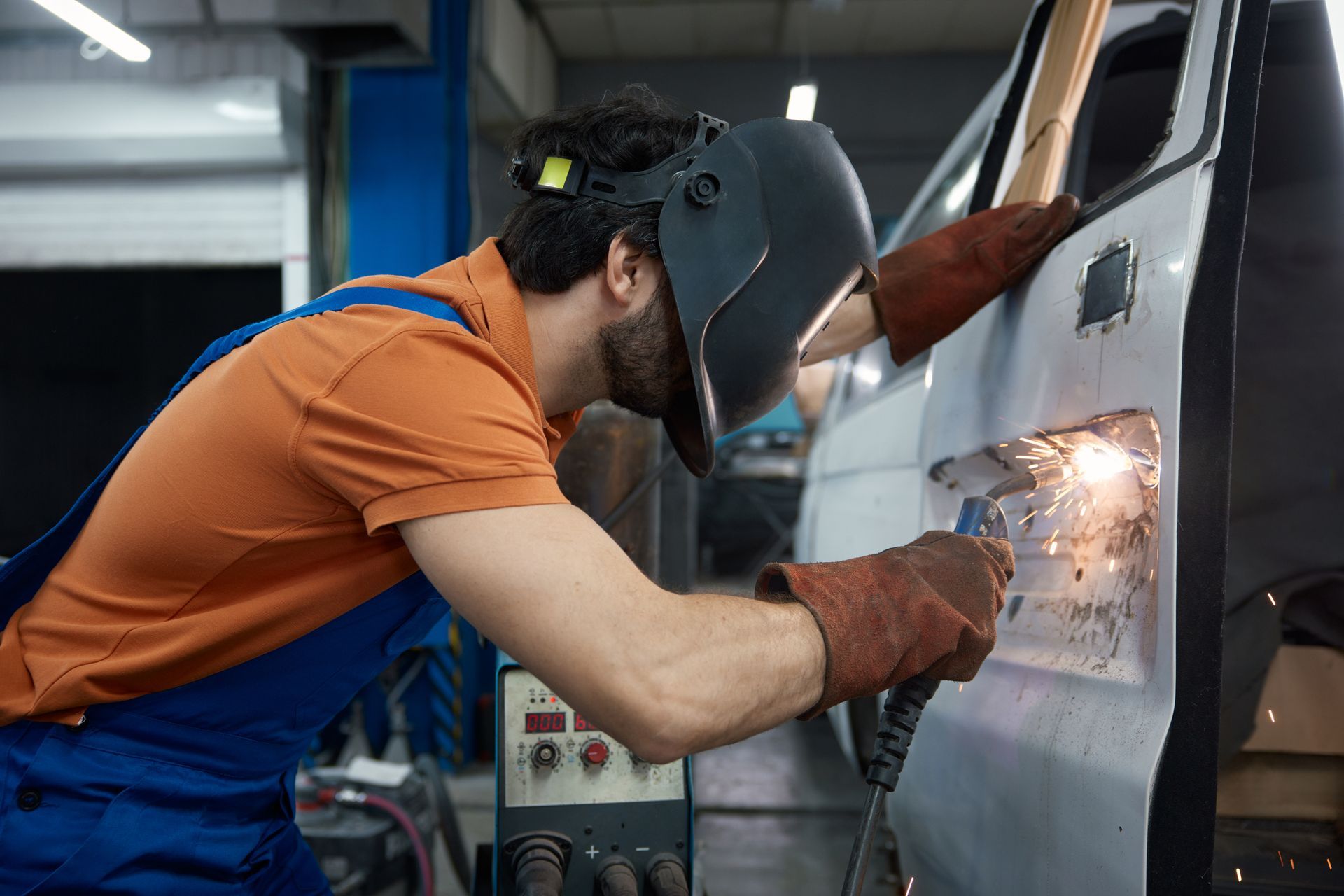 A welder in protective gear uses a torch to perform repair work on the metal body of a white van in a workshop.