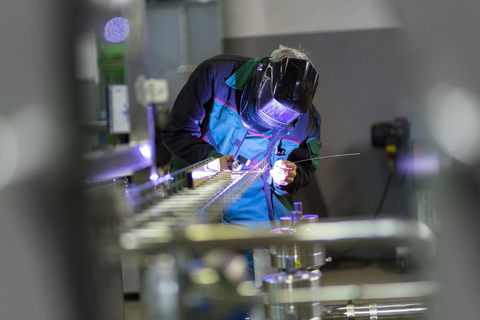 A person in a welding helmet and protective gear uses a TIG welder on a metal component in an industrial setting.