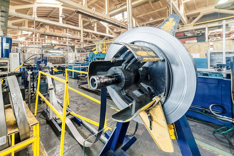 A metal coil unwinding on an industrial machine in a large factory with yellow safety railings.