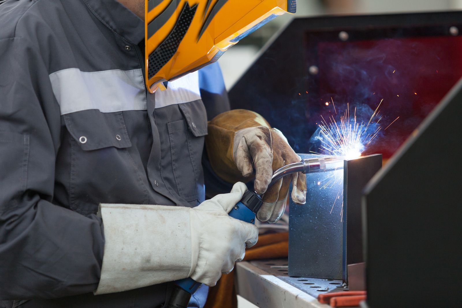 A welder in protective gear uses a torch to join metal pieces, creating bright sparks in a workshop.