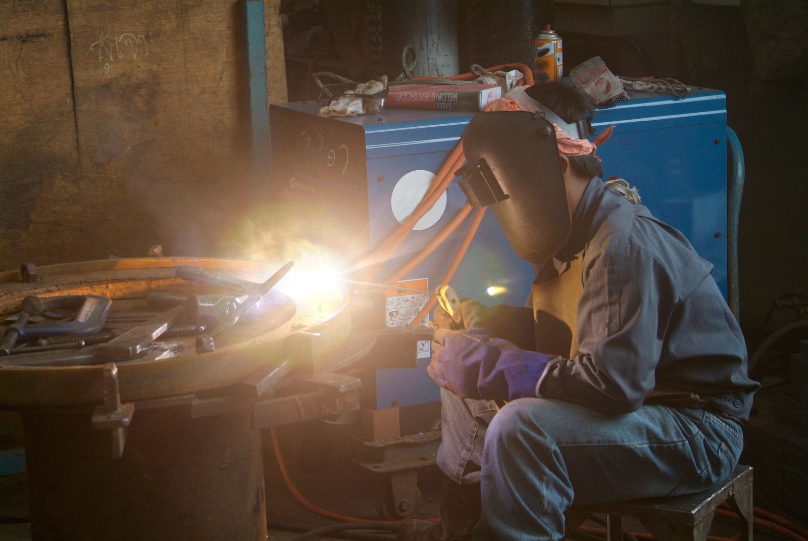 A person wearing a welding helmet and protective gloves works on metal, producing a bright glow in a workshop.