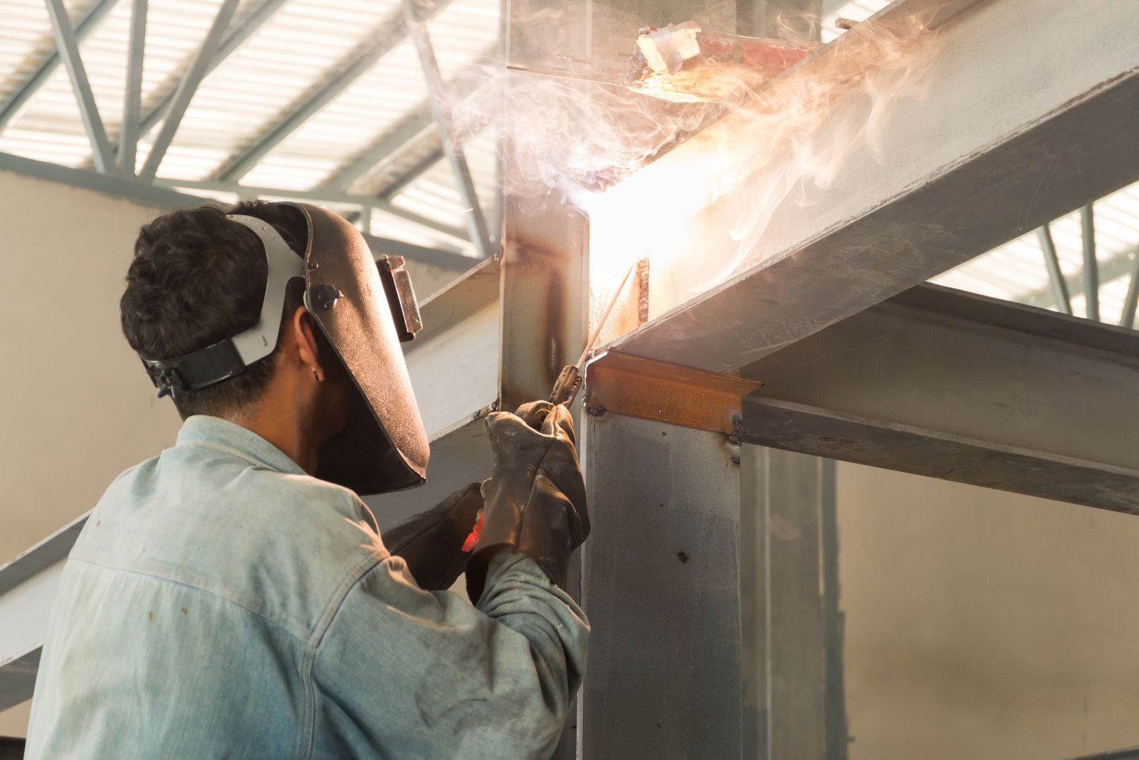 A worker wearing a welding helmet uses an arc welder on a metal structural beam in an industrial setting.