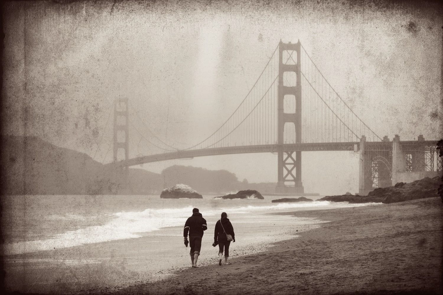 Two people walking on a beach with a bridge in the background