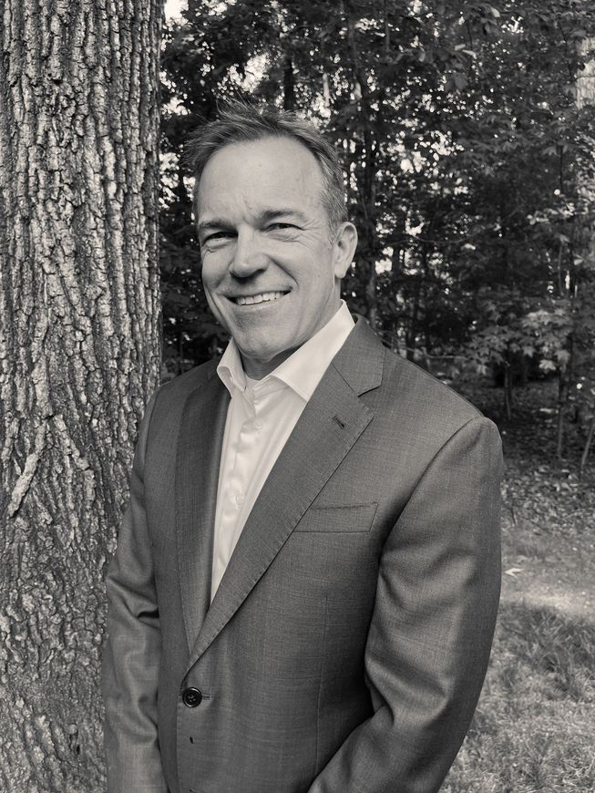 A man in a suit and white shirt is standing in front of a tree.