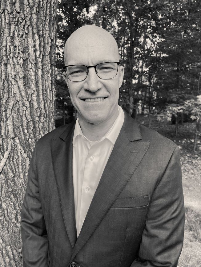 A man in a suit and glasses is standing in front of a tree.