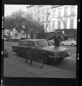A black and white photo of a man standing next to a car that says kodak extra 100