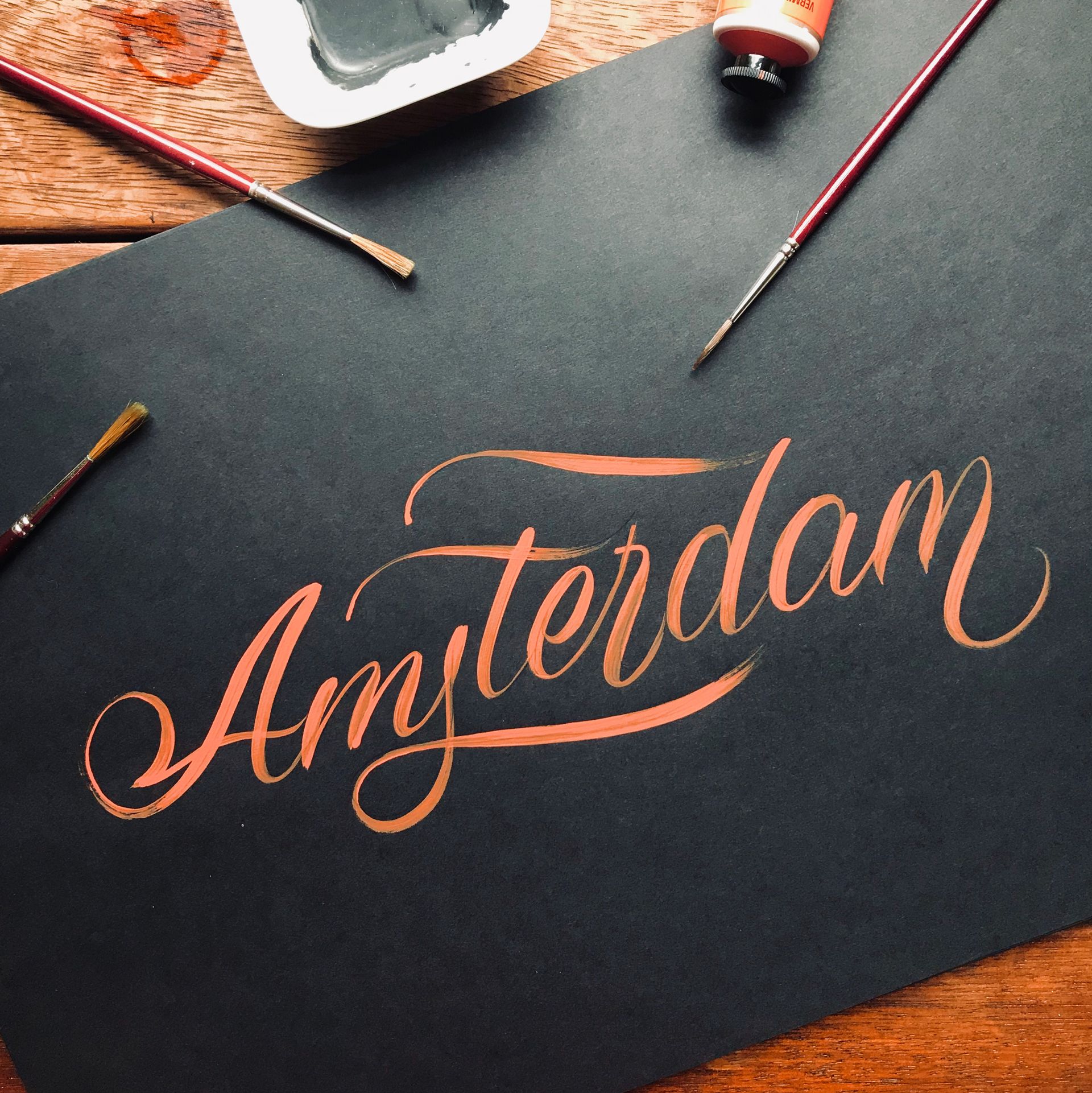 'Amsterdam' brush calligraphy written with orange acrylic paint over a black cardboard.
