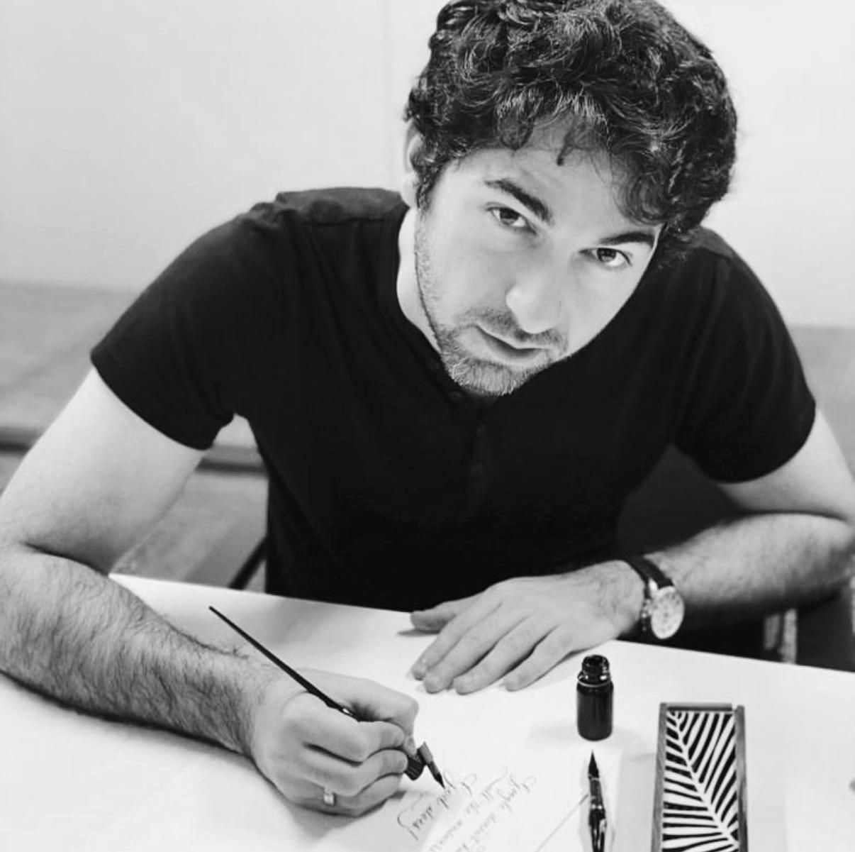 Person sketching at a desk with pen and paper, wearing a black shirt in a black-and-white photo