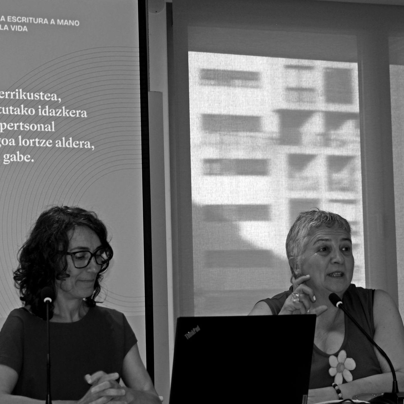 Two speakers at a panel table, one talking into a microphone with a laptop in front, black-and-white.
