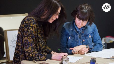 Portrait image of Maria Montes and a student of her at a desk reviewing some calligraphy writing.