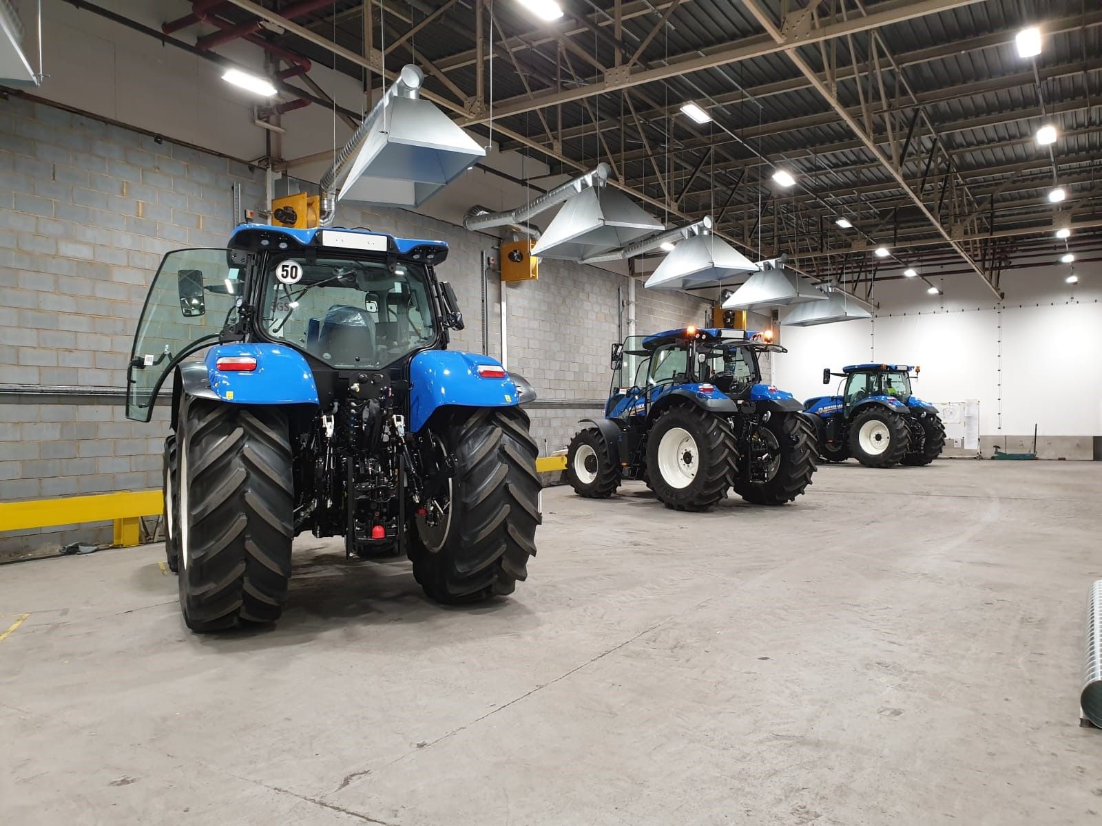 Blue tractors parked inside a warehouse with ventilation hoods overhead.