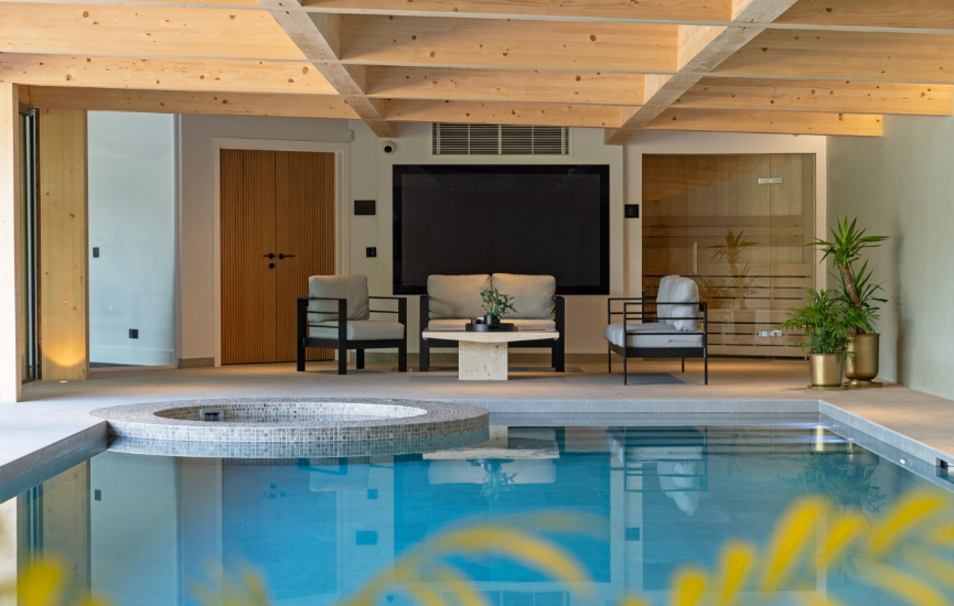 Indoor pool with seating area. Wooden beams, sauna, and jacuzzi. Blue water reflects ceiling.