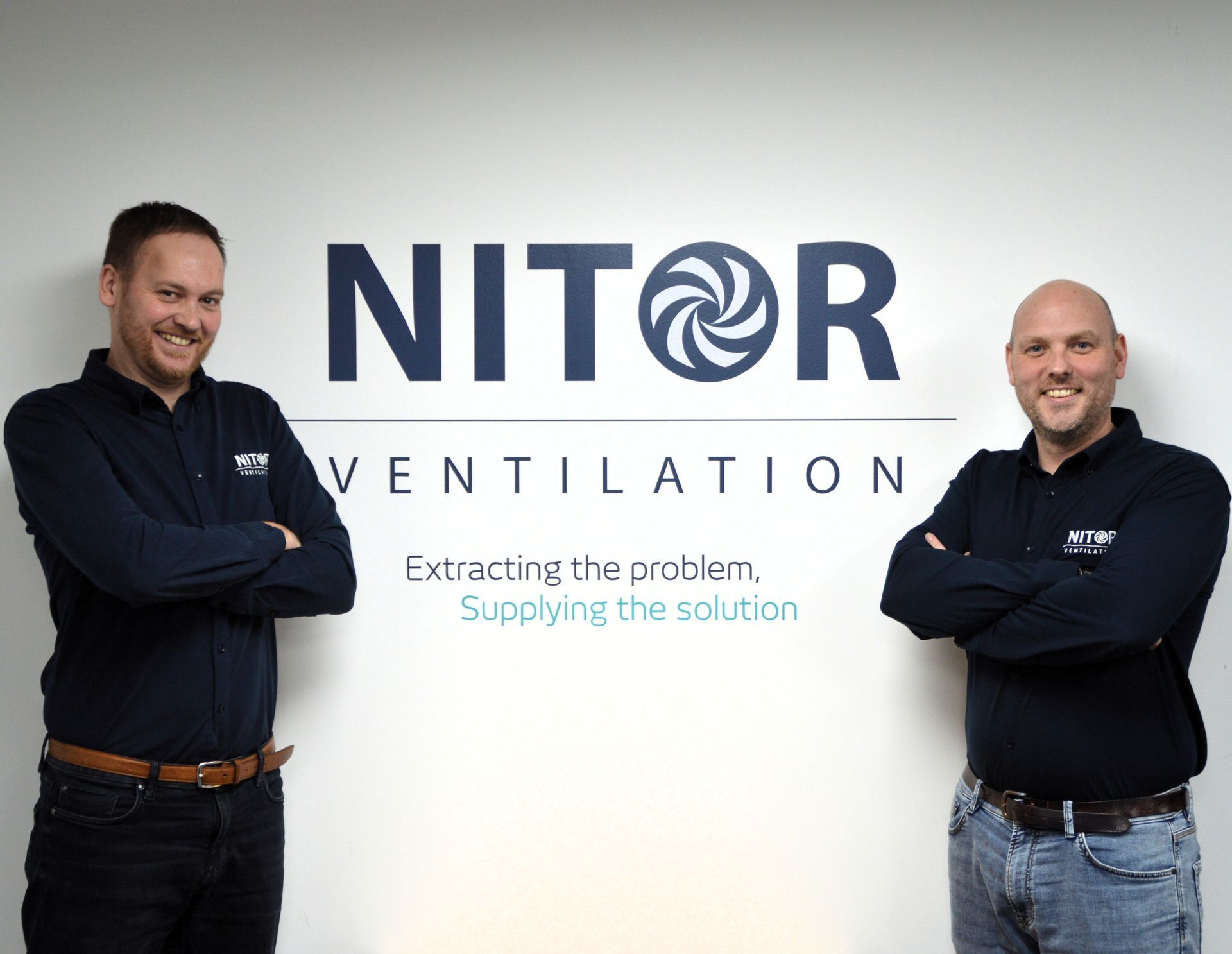 Two men in navy shirts stand in front of a wall with a logo for NITOR VENTILATION, arms crossed, smiling.