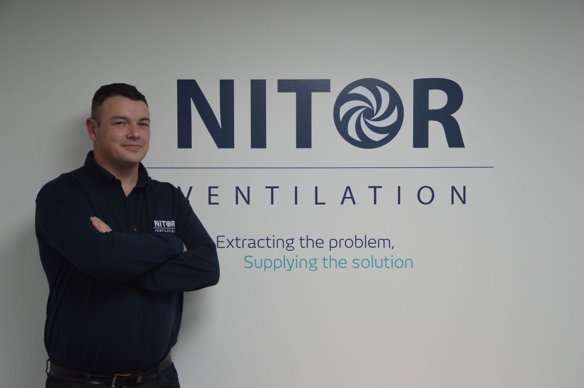 Man in blue shirt, arms crossed, standing in front of a 