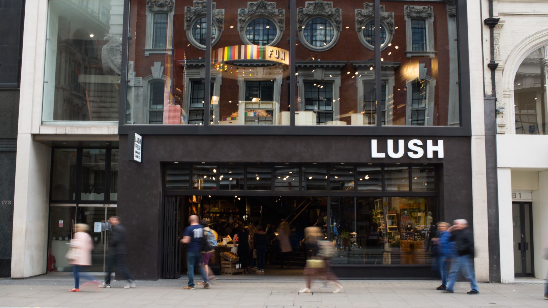Exterior of a Lush store with blurred people walking by. Black facade, large windows, and a gold light fixture.
