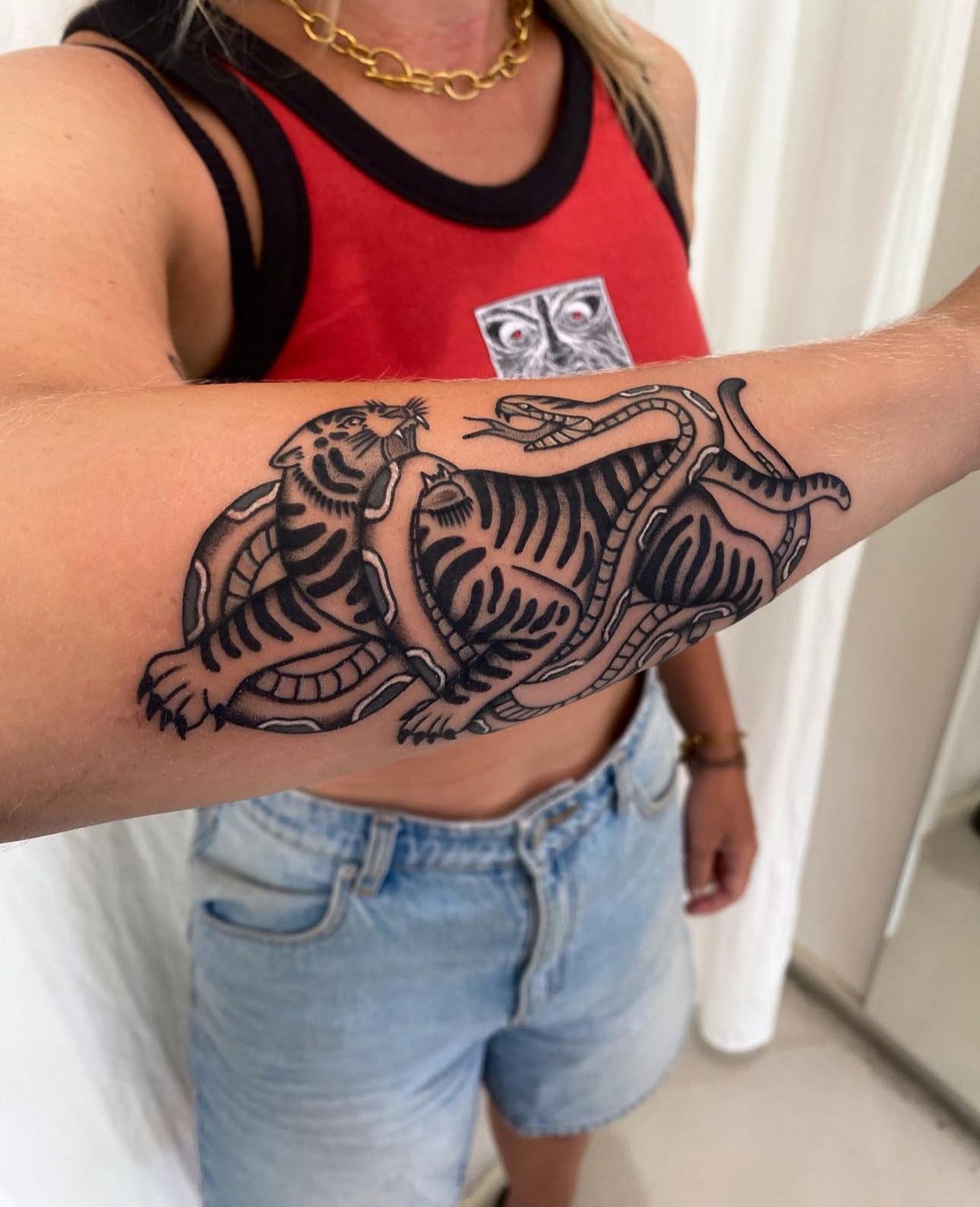 Person with a tattoo of two tigers in combat on their forearm; wearing red top and denim shorts.