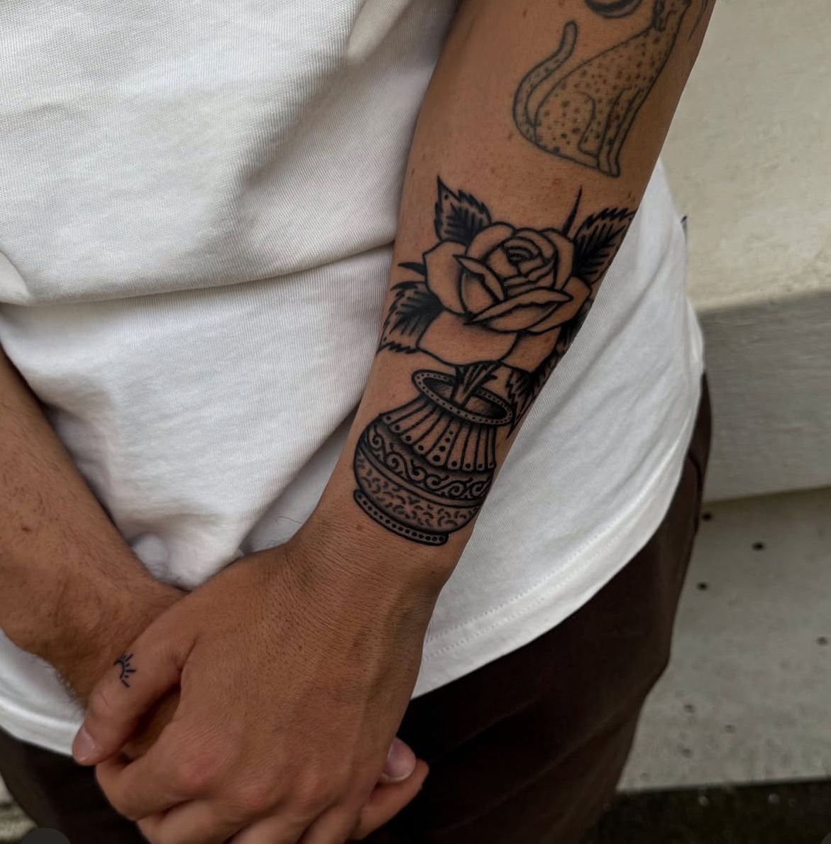 Person with forearm tattoo of a rose in a decorative vase. Black ink on white shirt.