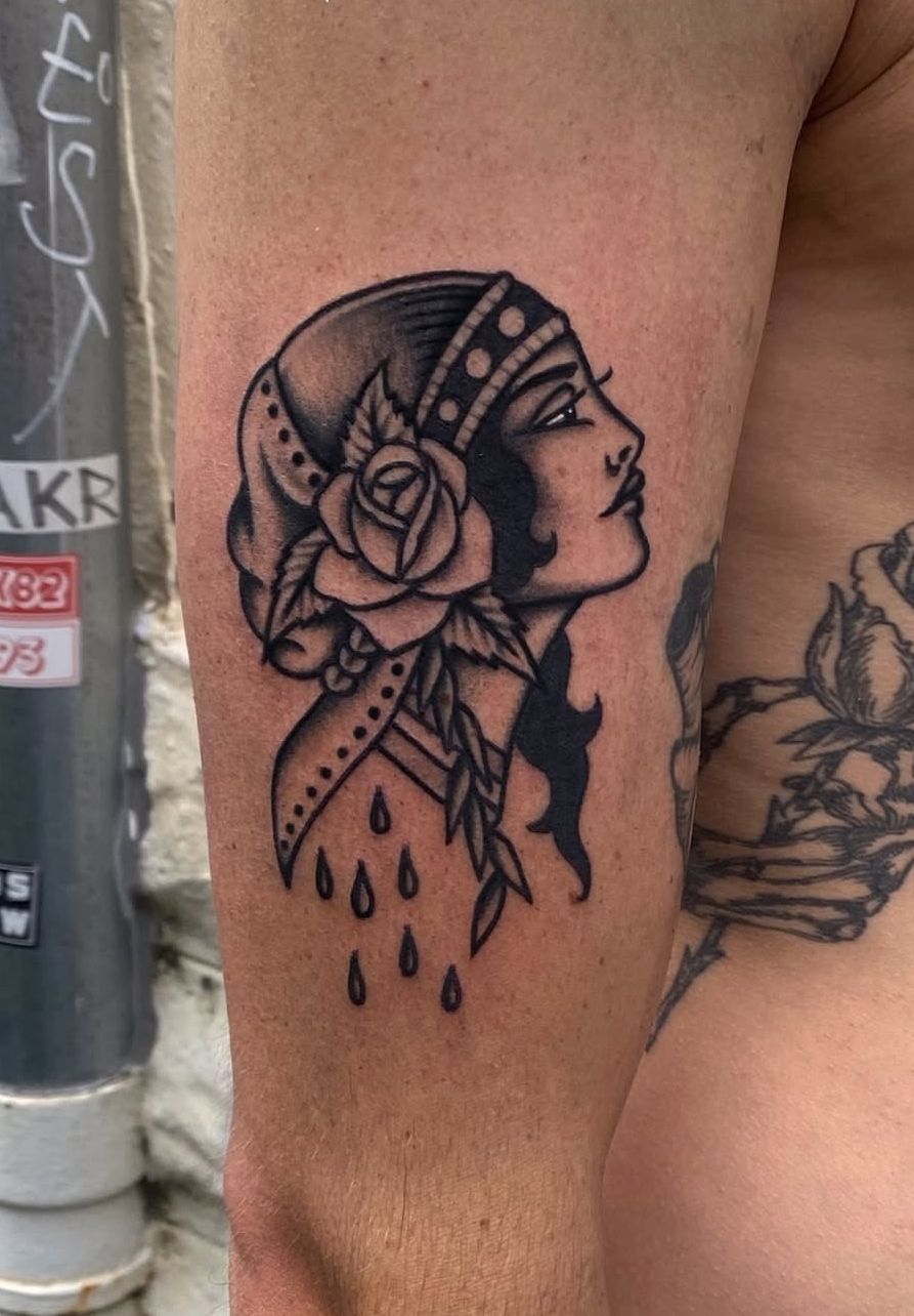 Black and white tattoo of a woman's profile wearing a headscarf, a rose, and tear drops, on a person's arm.