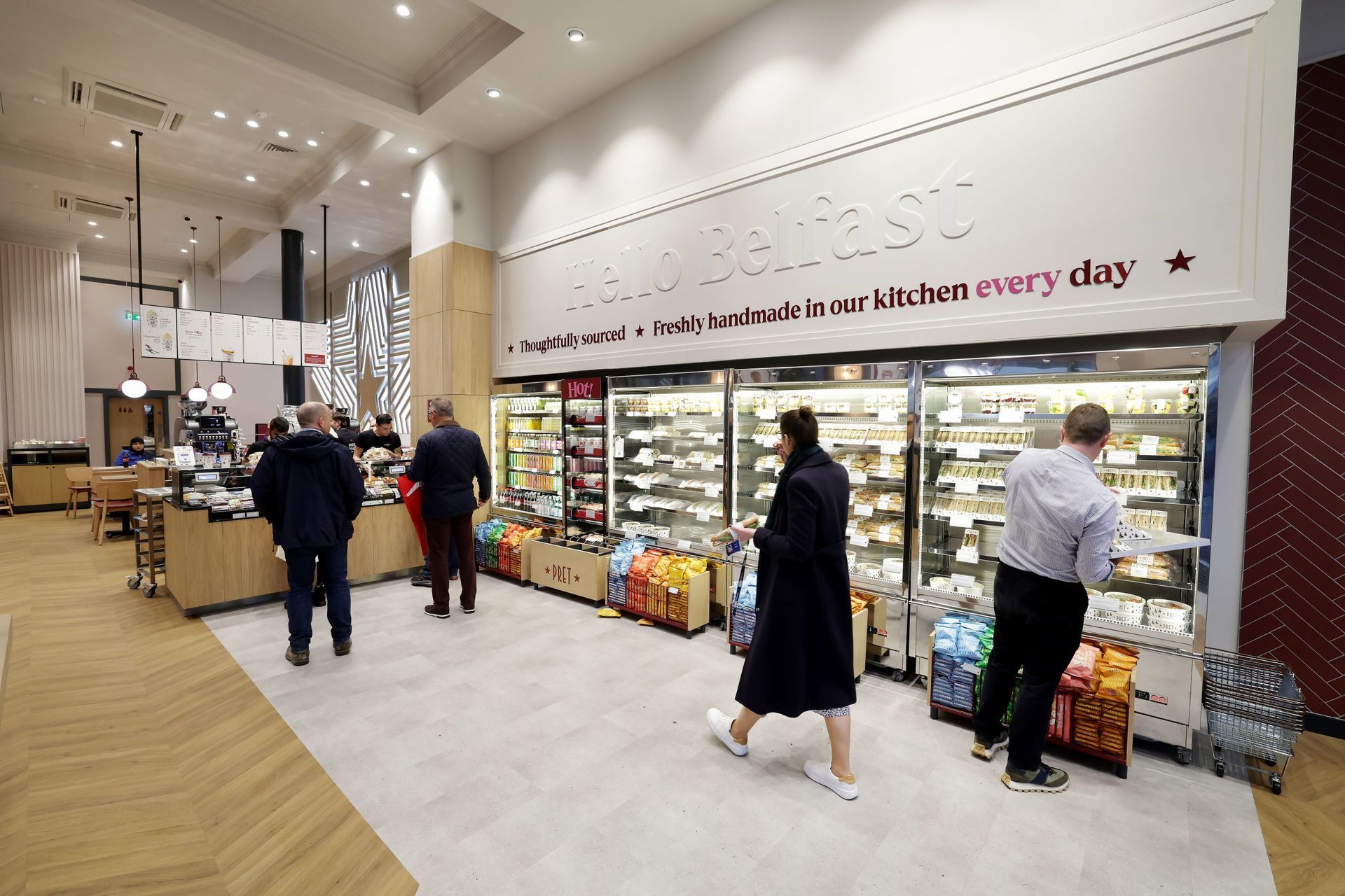 People browse inside a well-lit food store. Refrigerated shelves display food. Wooden counters, and a quote are on the wall.