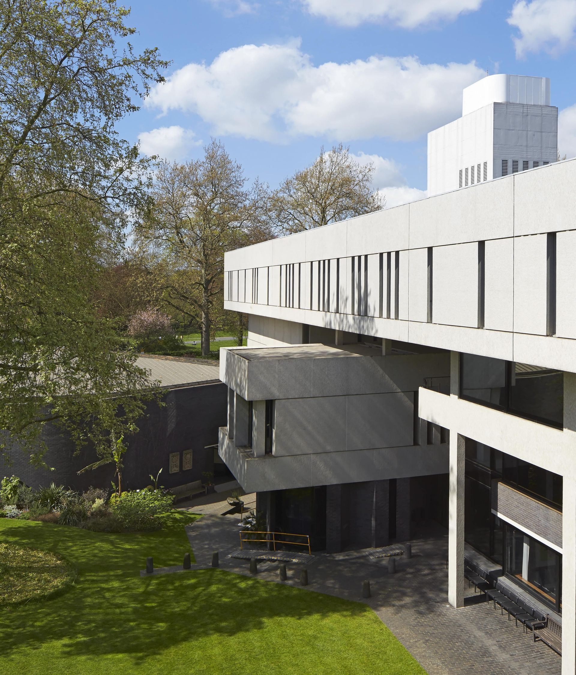 Modern concrete building with rectangular windows under blue sky, surrounded by green trees and grass.