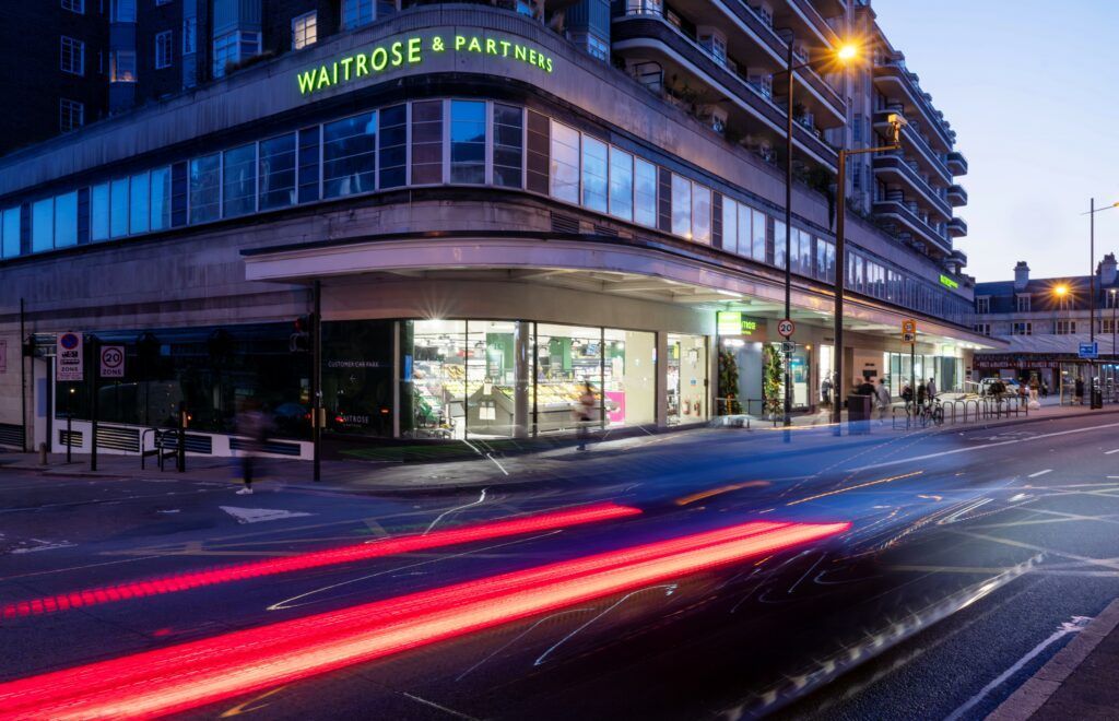 Waitrose store at dusk, with bright interior lights and passing car light trails.
