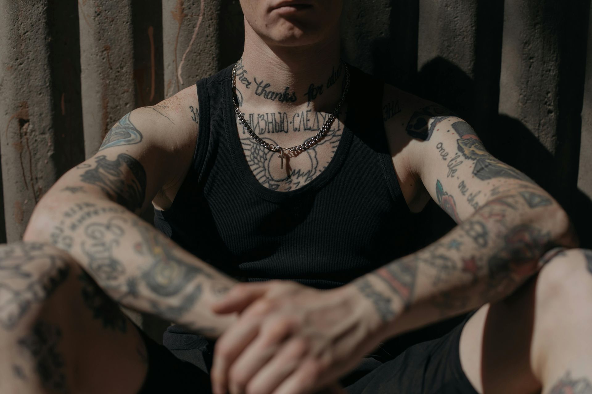 Man with tattoos sits, arms crossed, wearing black tank top, against corrugated metal.