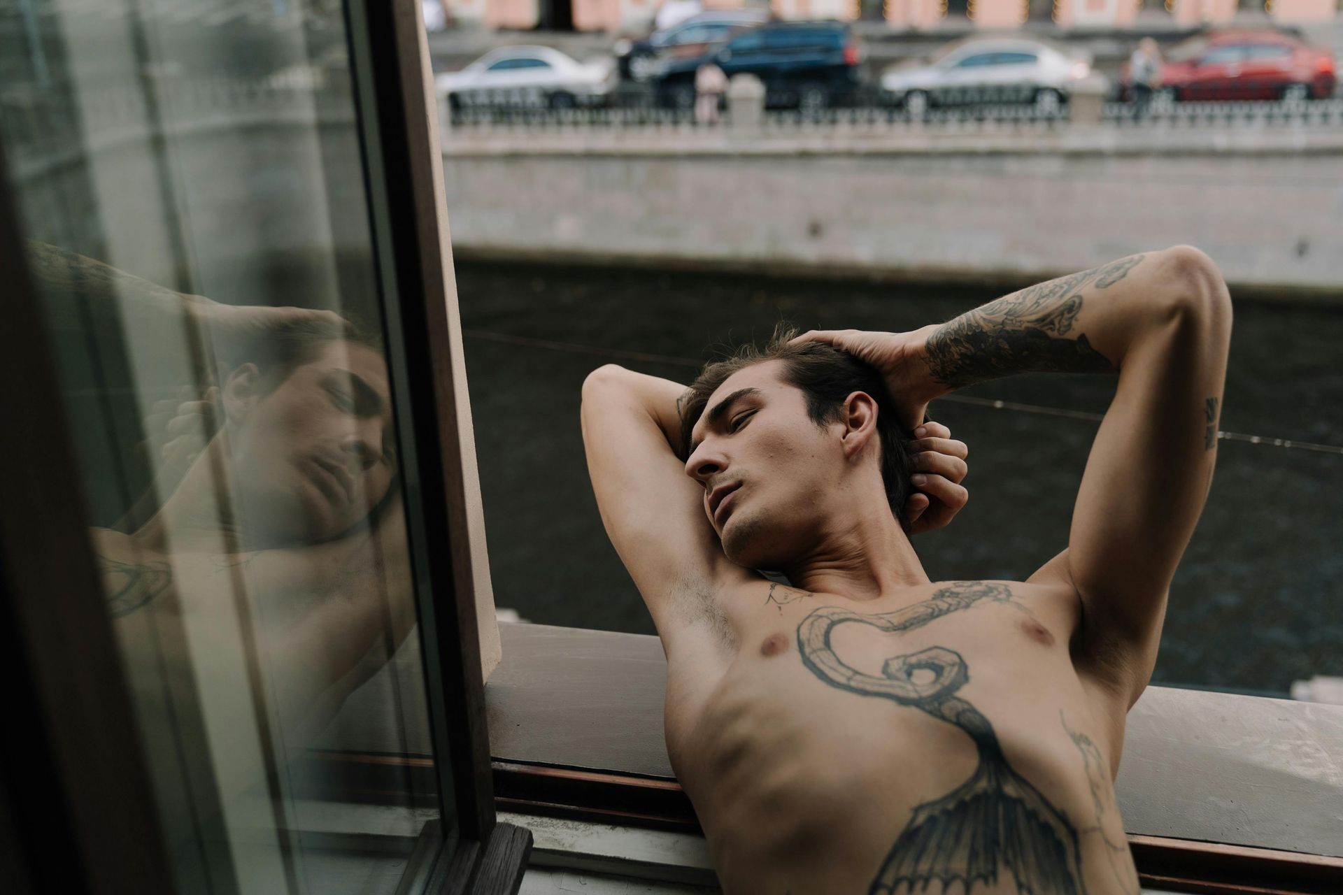 Man with tattoos relaxes, leaning against a window with reflection of him, overlooking a canal.