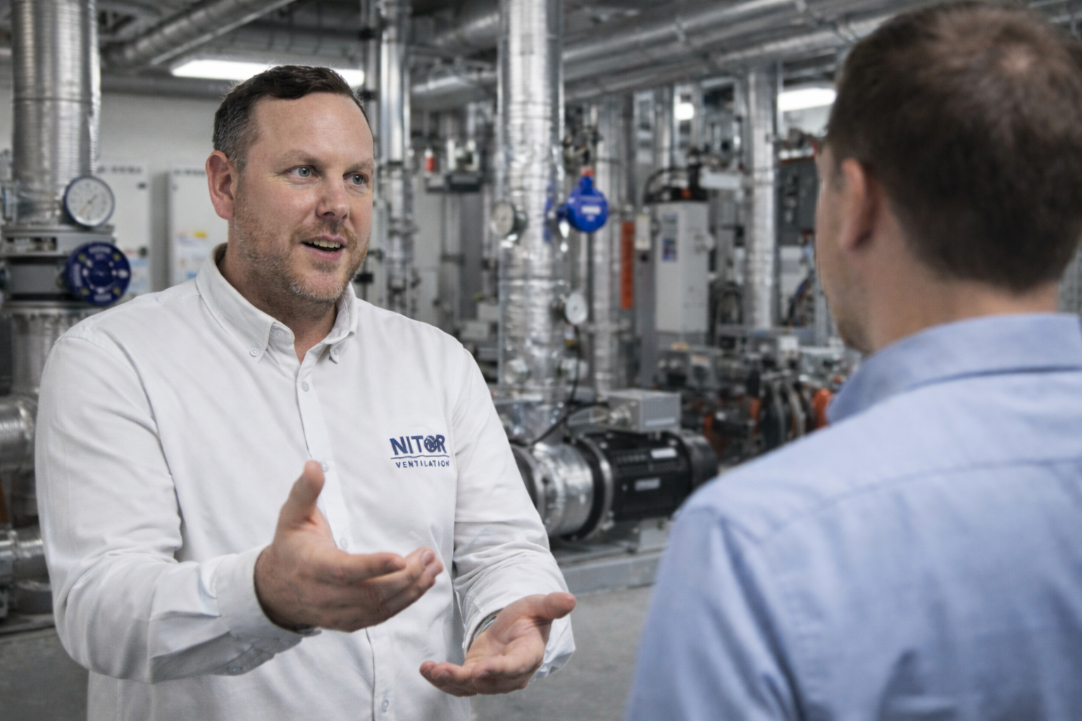 Man in white shirt gesturing, talking to another man in an industrial setting with machinery.