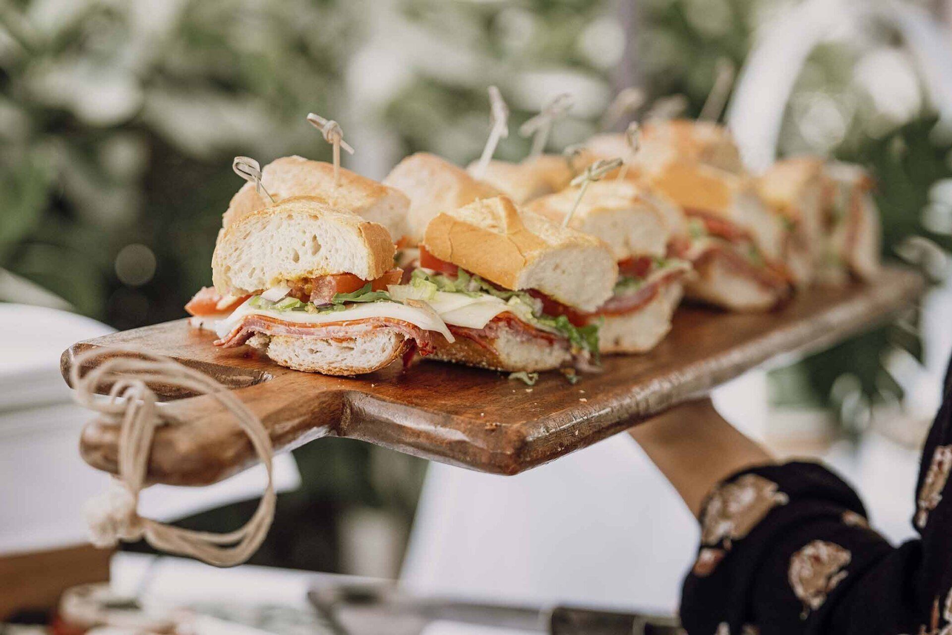 A person is holding a wooden cutting board with sandwiches on it.