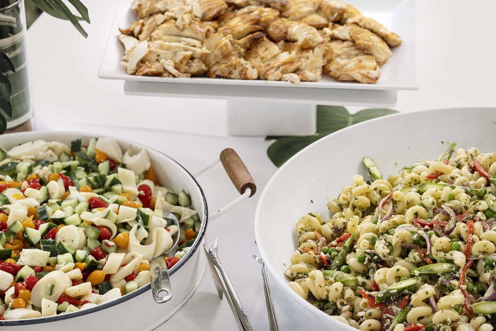 Three bowls of pasta salad are on a table next to a plate of chicken.