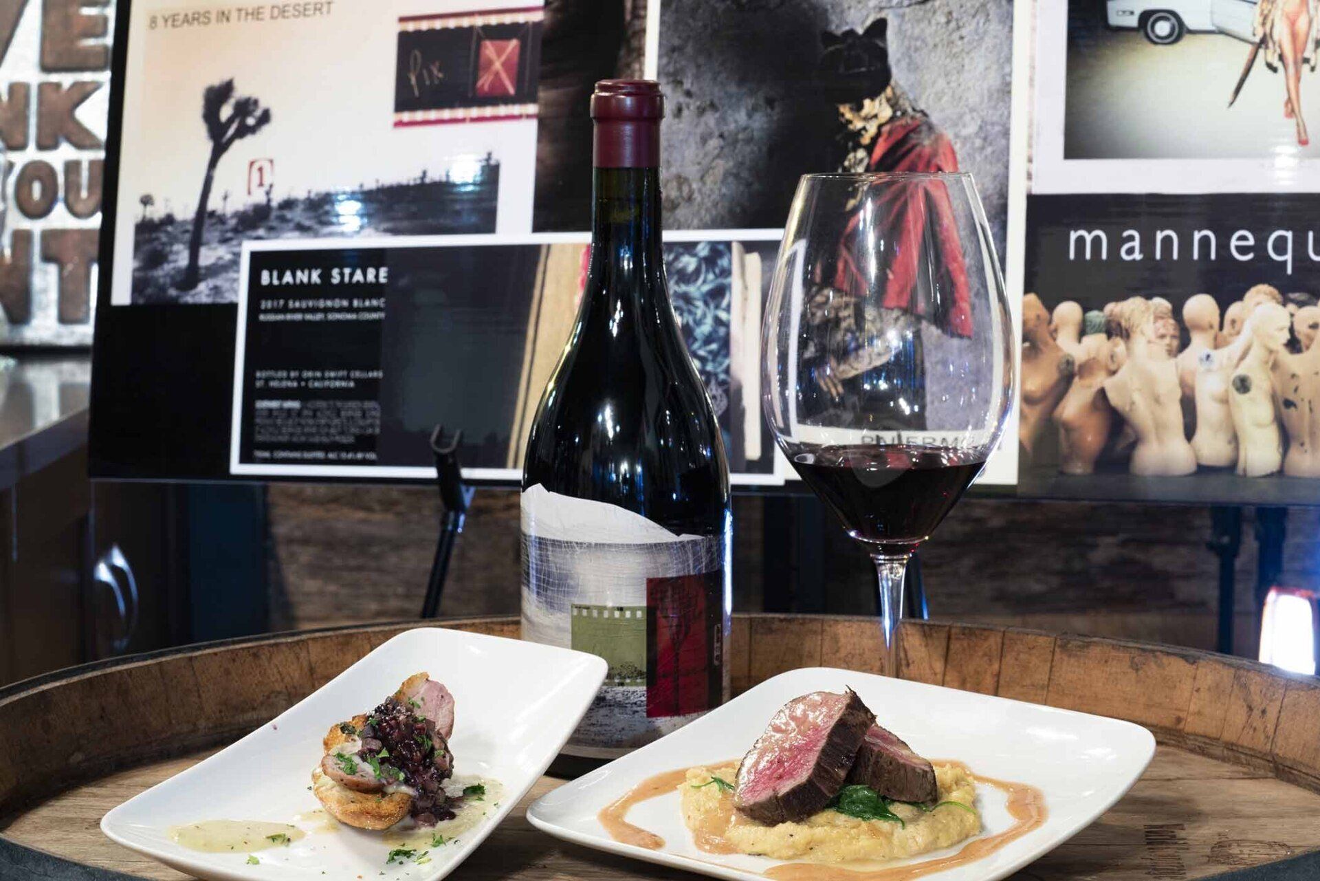 A bottle of wine and two plates of food are on a wooden barrel.