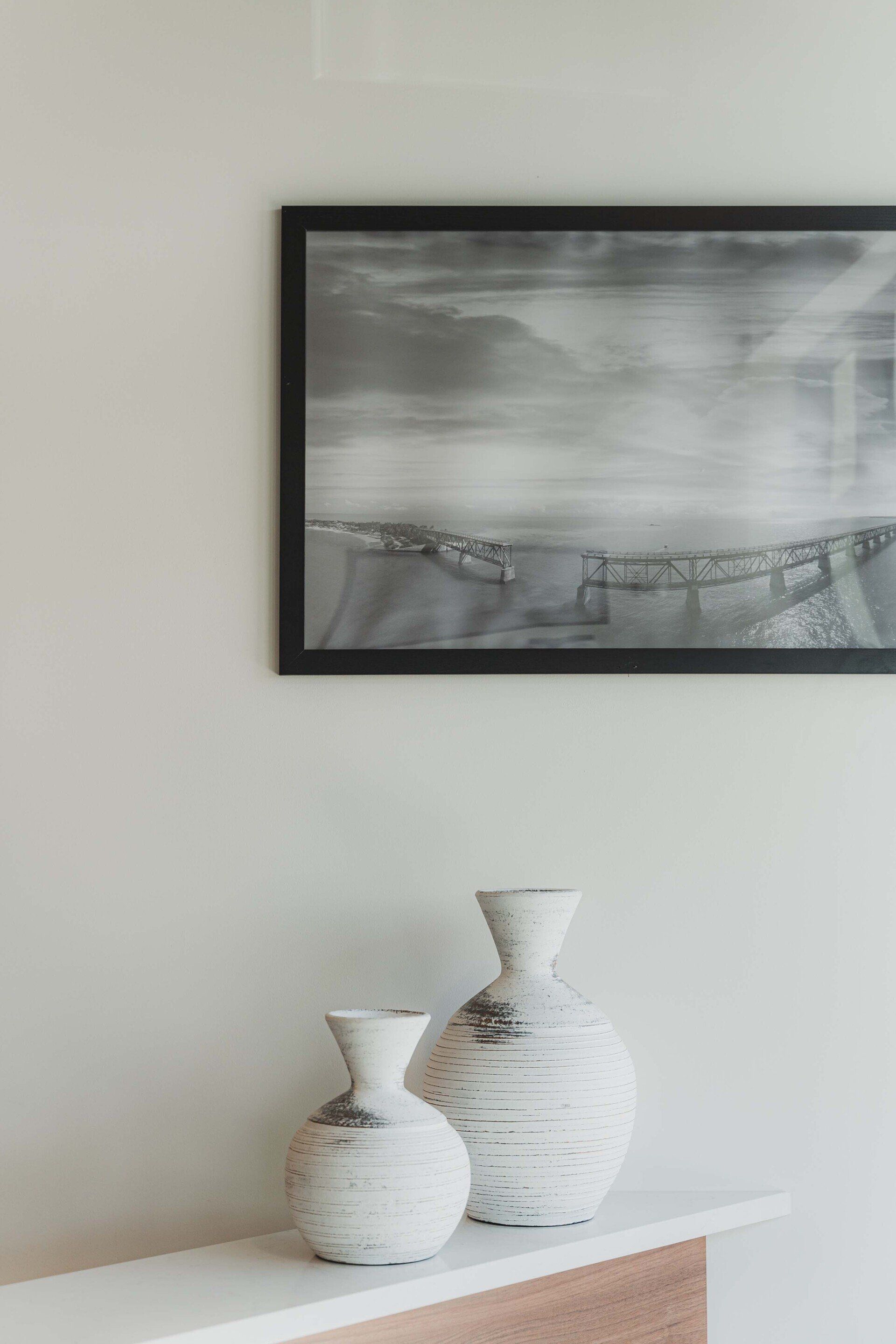 Two vases are sitting on a shelf under a painting on the wall.