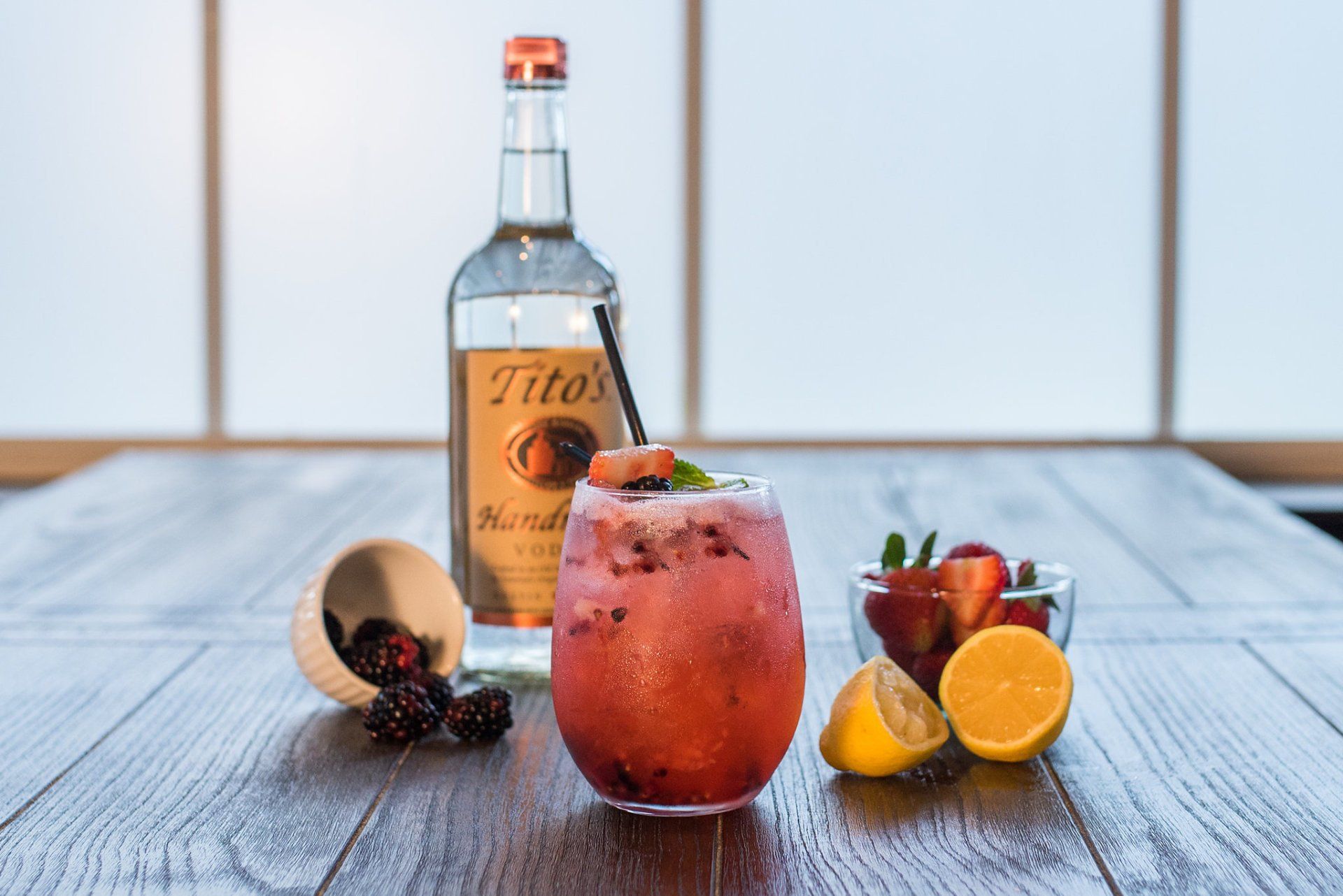 A bottle of tito 's vodka is sitting on a wooden table next to a glass of drink.