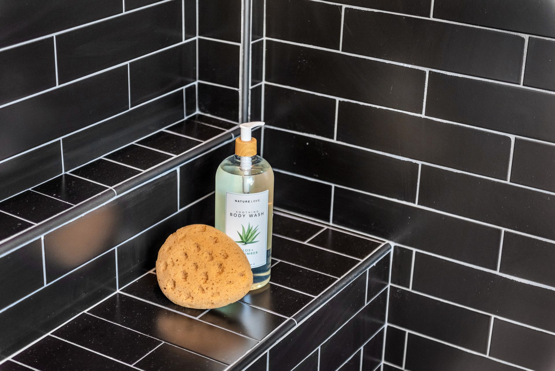 Black tiled shower with a shelf holding soap, sponge, and shower gel.