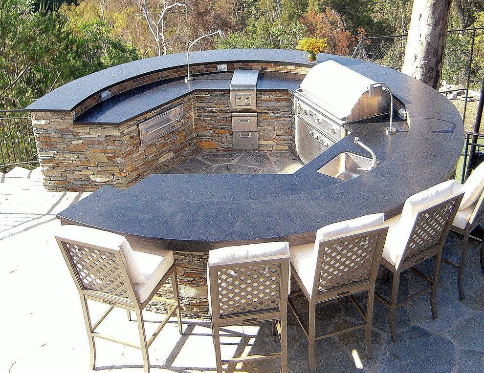 Outdoor curved stone-clad kitchen with a grill, sink, bar, and seating.