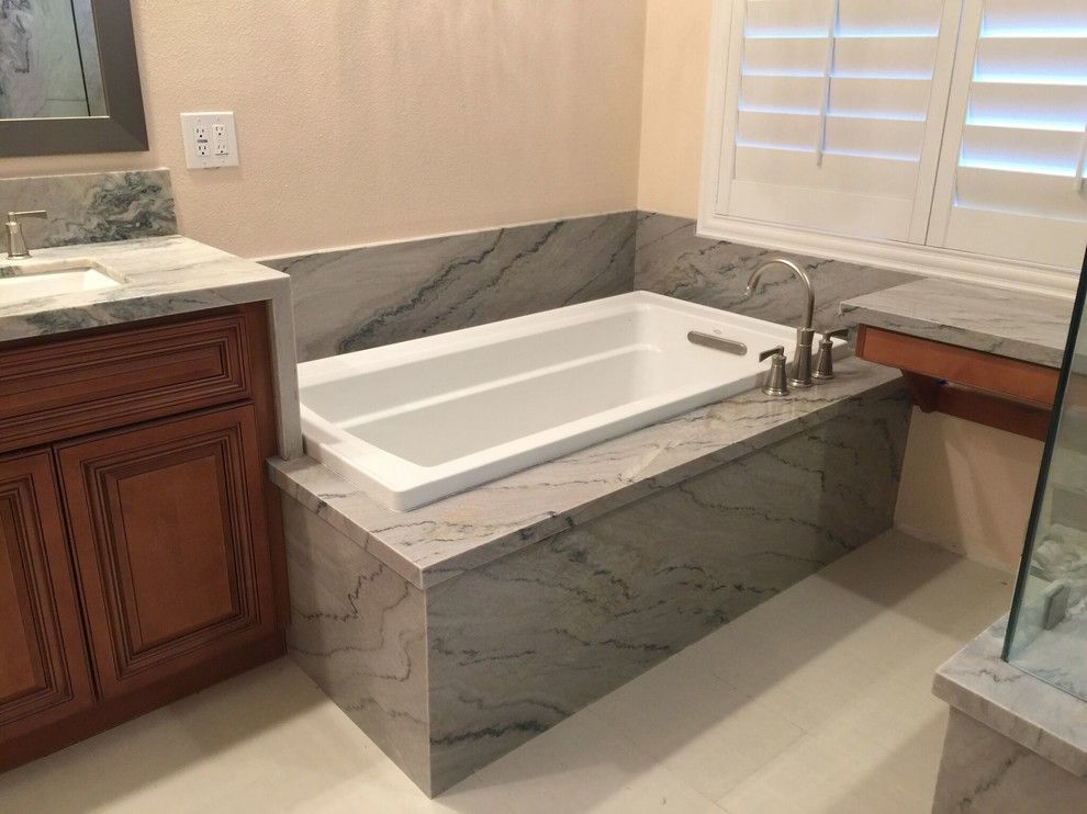 Bathroom with a white bathtub, gray marble surround, wooden vanity, and window with shutters.