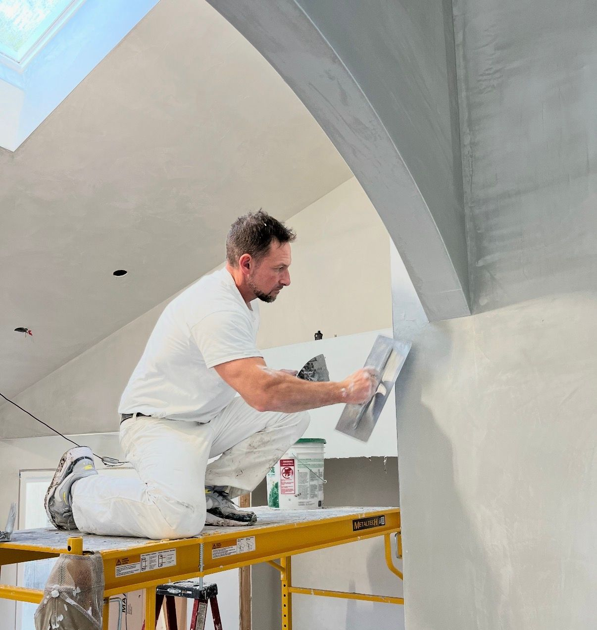 Interior of a house under construction; white walls, vaulted ceiling, orange ladder in foreground, construction materials on the floor.