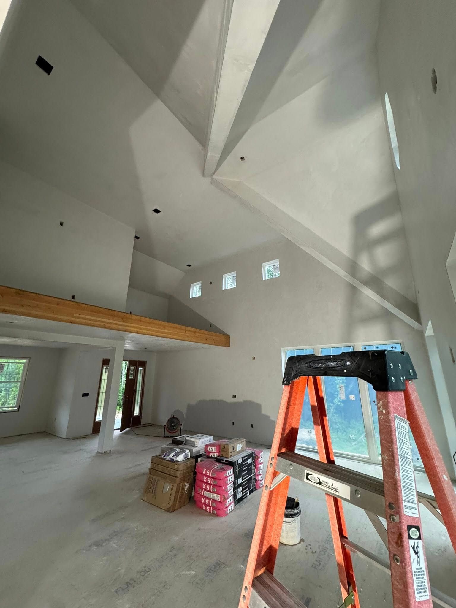 Interior of a house under construction; white walls, vaulted ceiling, orange ladder in foreground, construction materials on the floor.