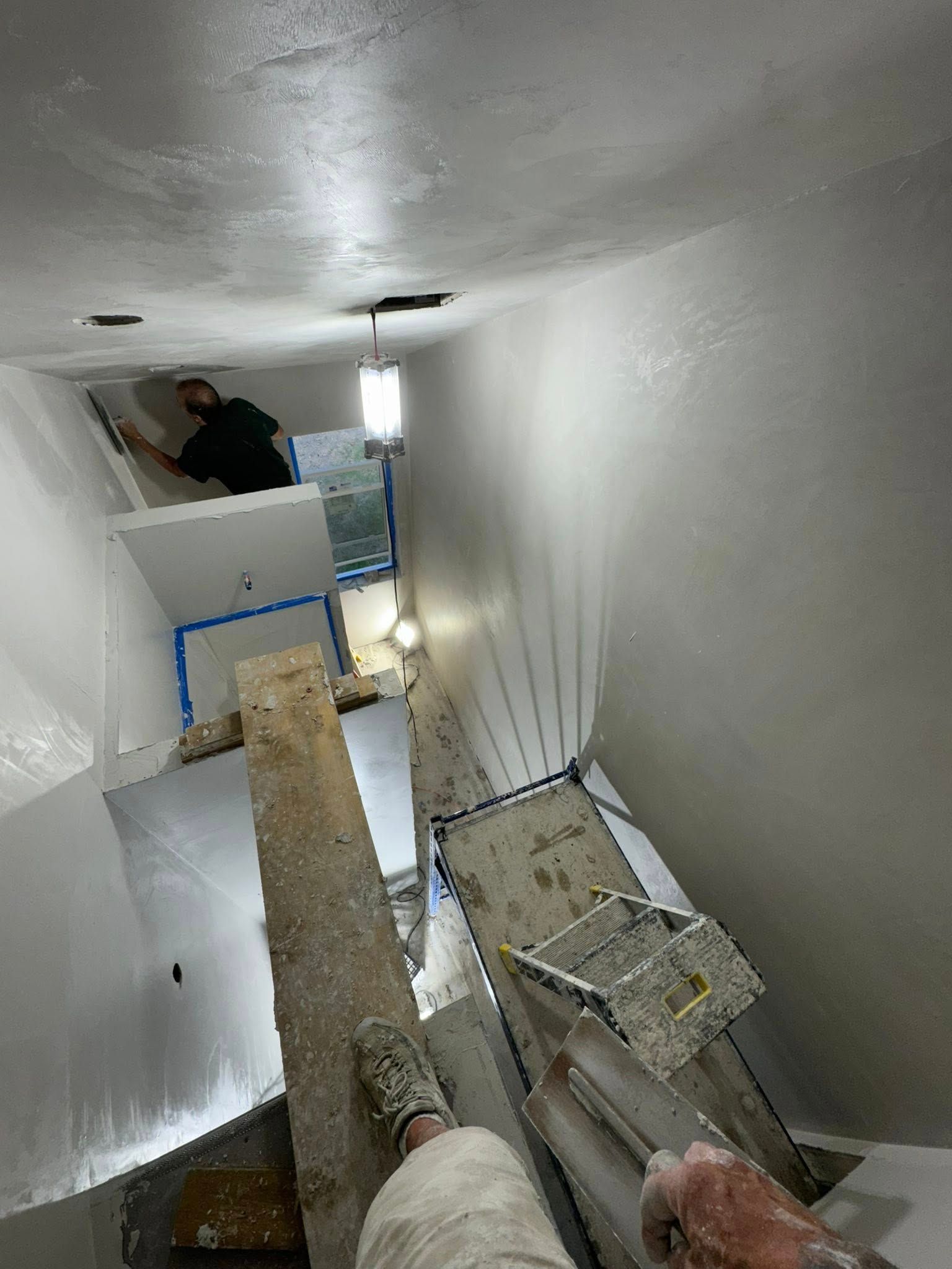 Person applies coating to white walls in a narrow space with scaffolding and overhead lights.
