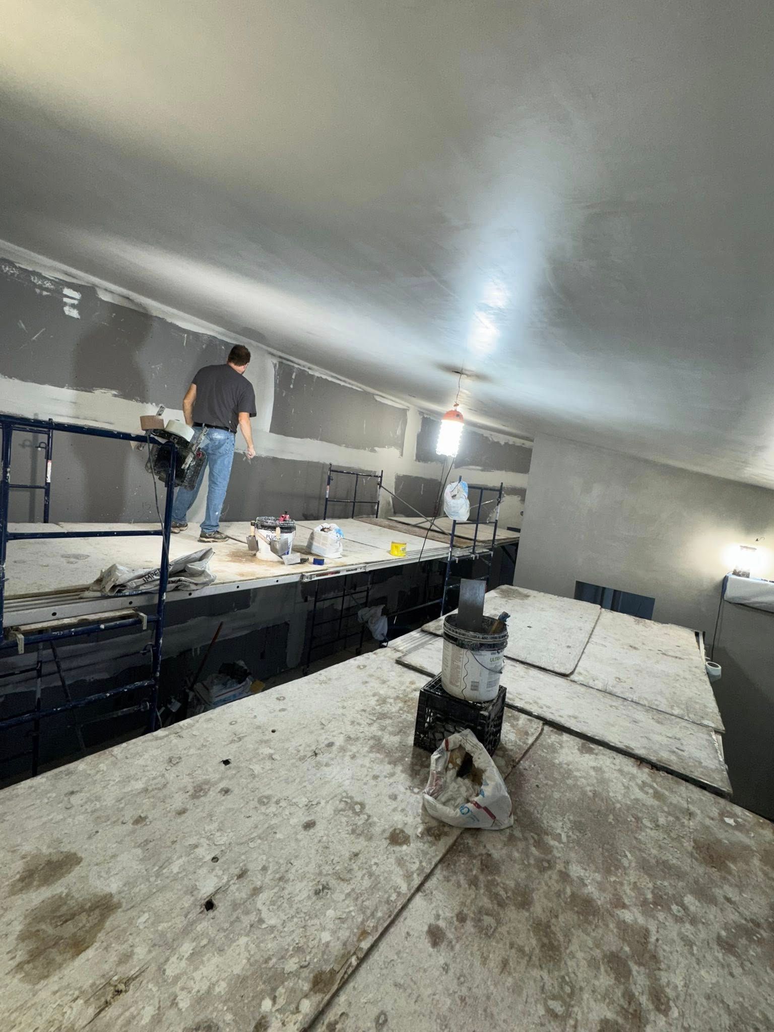 A person on a lift works on drywall in a construction setting. A person on a lift works on drywall in a construction setting.