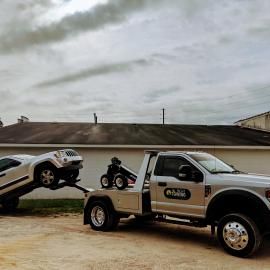 A white tow truck towing a white SUV, parked on a tan surface, cloudy sky backdrop.