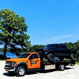 Orange tow truck carrying a black pickup truck on a sunny day near a body of water and trees.