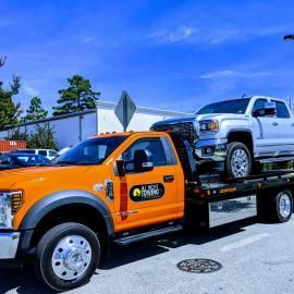Orange tow truck carrying a silver pickup truck on a sunny day.
