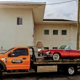 Orange tow truck with a red classic car on the flatbed, parked in front of a building.