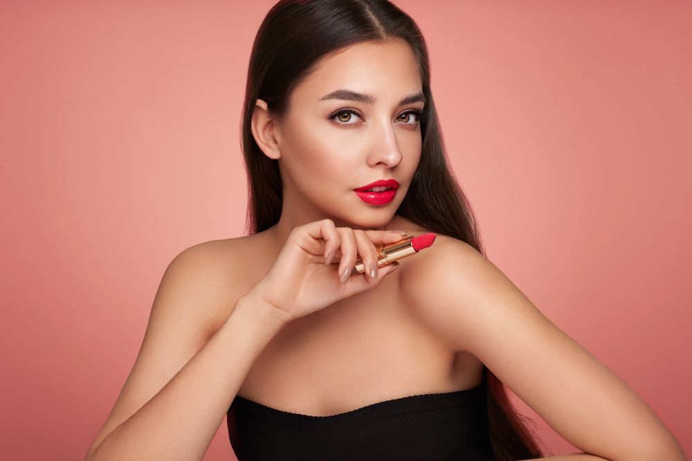 Una persona con cabello largo y oscuro sostiene un lápiz labial rojo sobre un fondo rosa liso
