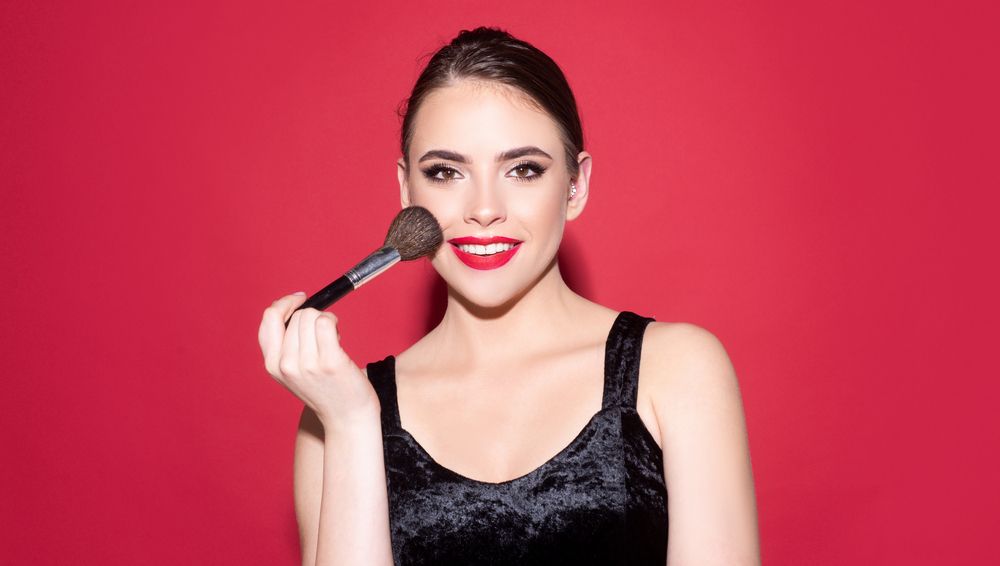 Una persona sonriente, vestida con una blusa negra, sostiene una brocha de maquillaje en su mejilla 