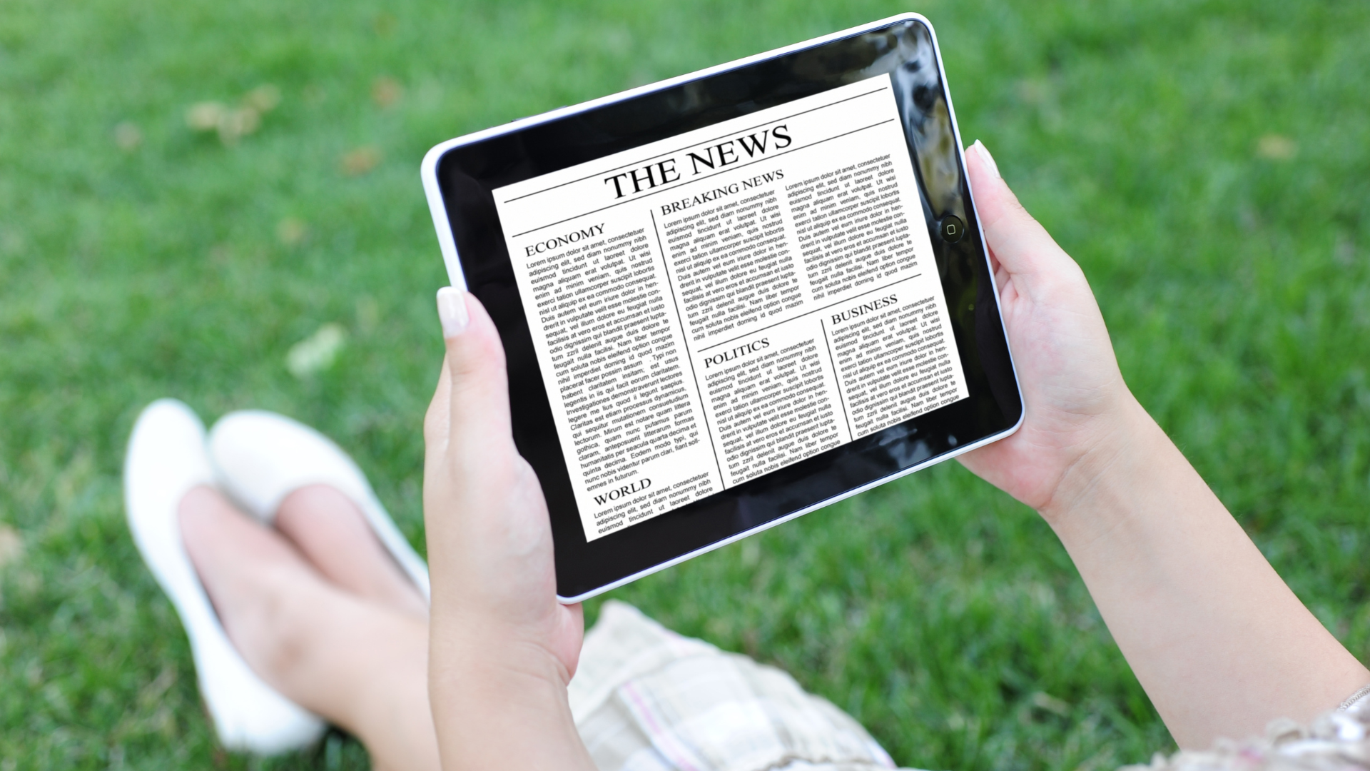 Person holding a tablet outdoors, displaying a newspaper on a grassy lawn.
