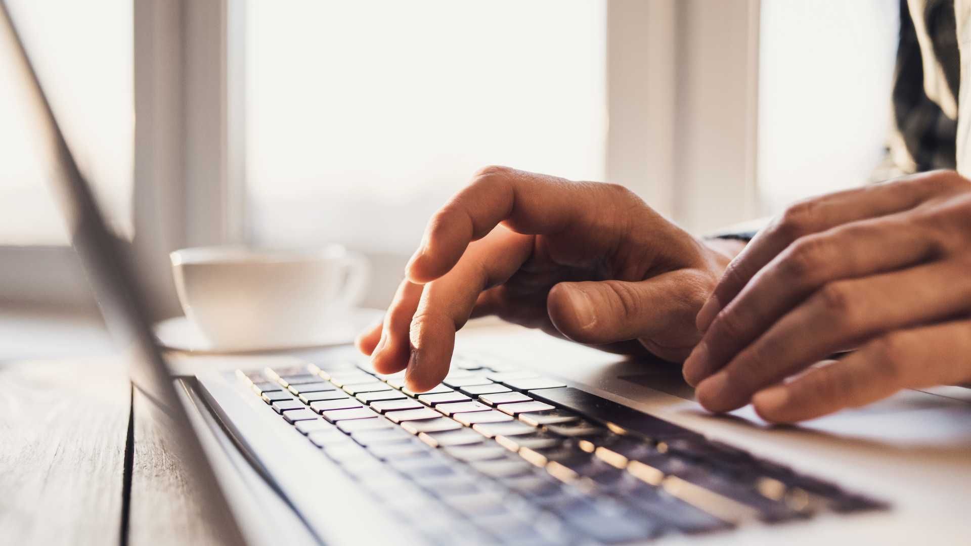 Hands typing on a laptop keyboard; a cup sits in the background near a window.